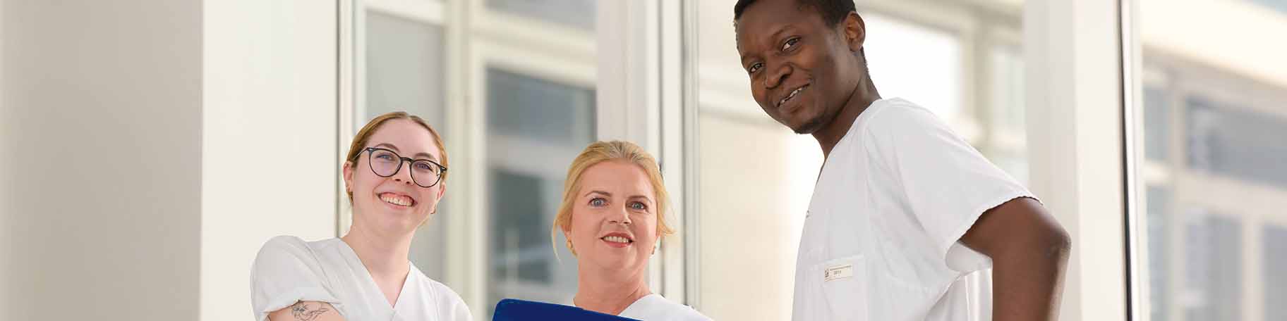 Drei Pflegekräfte stehen im Gang mit einem Klemmbrett und schauen lachend in die Kamera